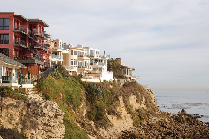 A row of colorful houses with balconies sits atop a steep cliff overlooking the blue ocean in a photo taken in Corona del Mar, California during the summer afternoon.