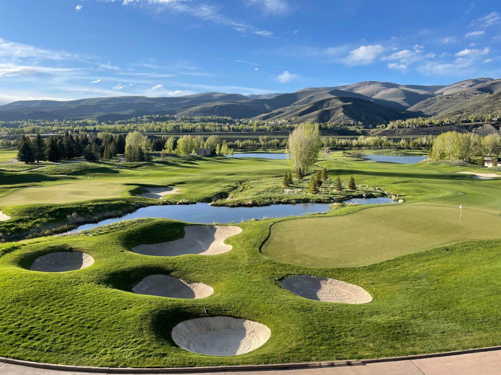 Green grass at a golf course near green trees and mountains under blue sky during daytime