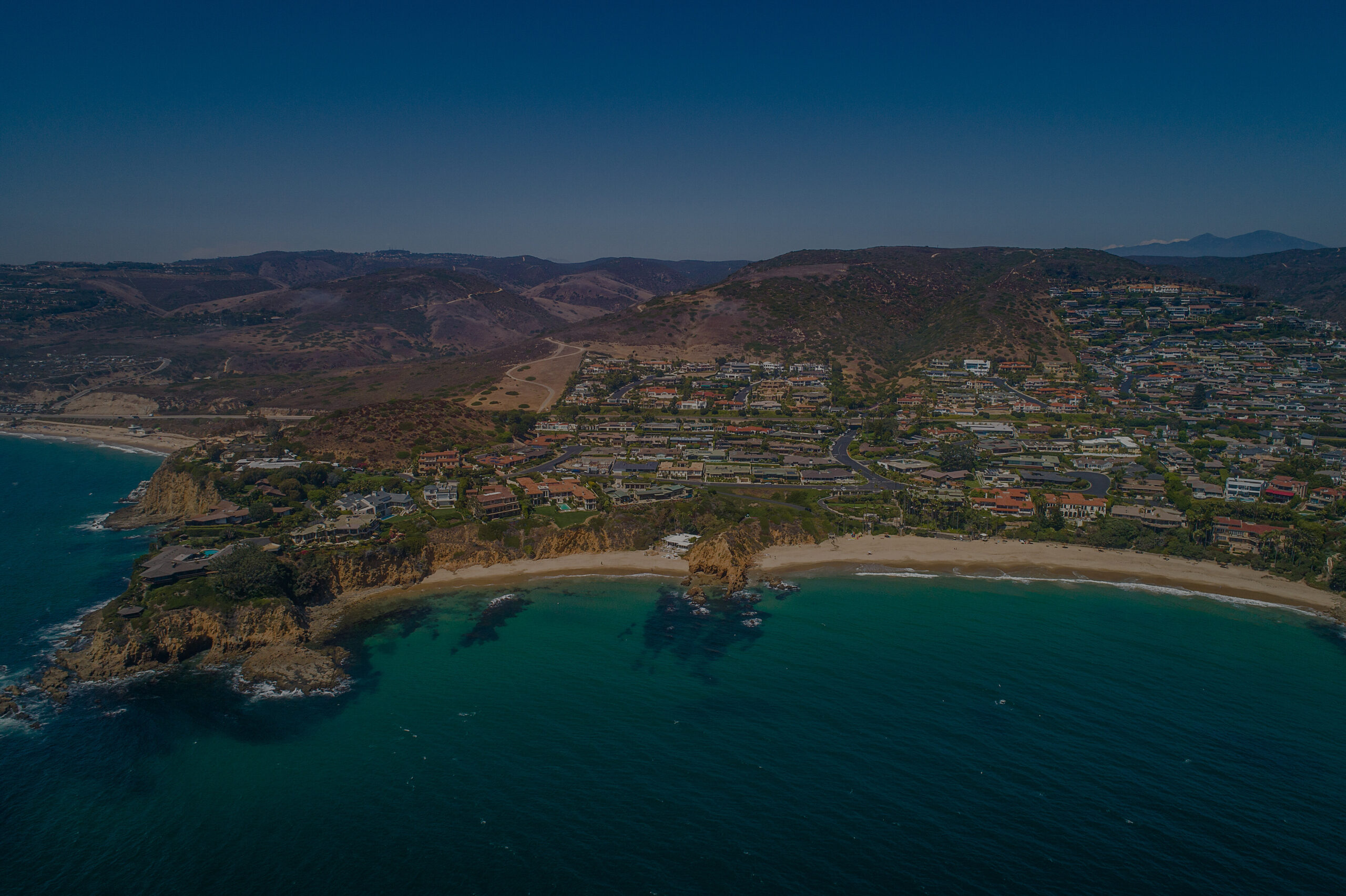 Drone view of luxury blufftop homes in Coastal Orange County overlooking the Pacific Ocean with crashing waves below, highlighting the exclusive real estate market of Newport Beach, Laguna Beach, and Corona del Mar.
