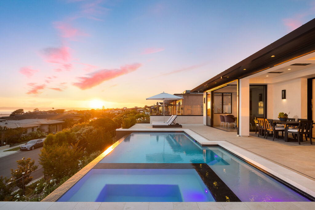 A breathtaking sunset view from the infinity pool and patio of a luxury home in Corona Del Mar sold by the Stavros Group. The outdoor space features sleek modern design, with a pool that appears to merge with the horizon, offering unobstructed views of the Pacific Ocean. This property exemplifies the indoor-outdoor living lifestyle that is highly sought after in Corona Del Mar luxury real estate.