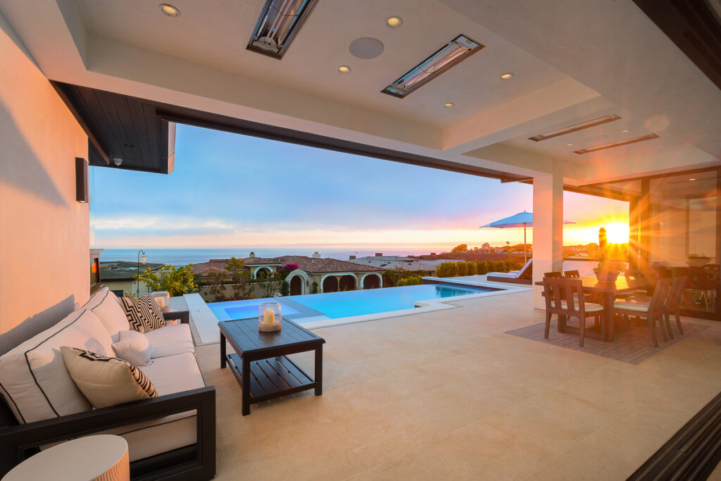 A luxurious California room with open-air access to a patio, offering seamless indoor-outdoor living at a high-end Corona Del Mar property sold by top Realtors, the Stavros Group. The room is designed for comfort and style, featuring cozy seating areas and large glass doors that frame stunning sunset views over the Pacific Ocean.